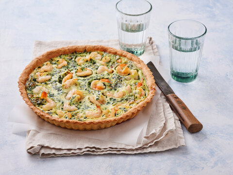 A quiche filled with shrimp and spinach sits on a linen cloth beside two glasses of water. The setting is a simple kitchen table. Light shines softly on the dish