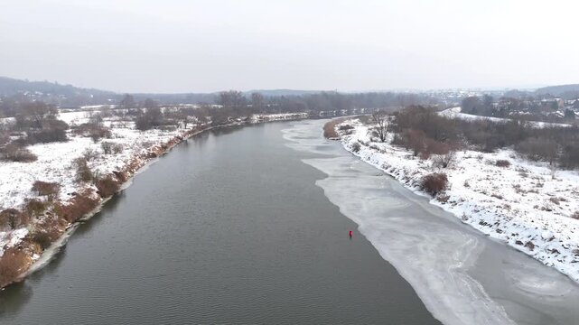 Aerial view of a tranquil river winding through a snowy landscape, showcasing ice formations along the banks and patches of snow-covered ground in the surrounding area