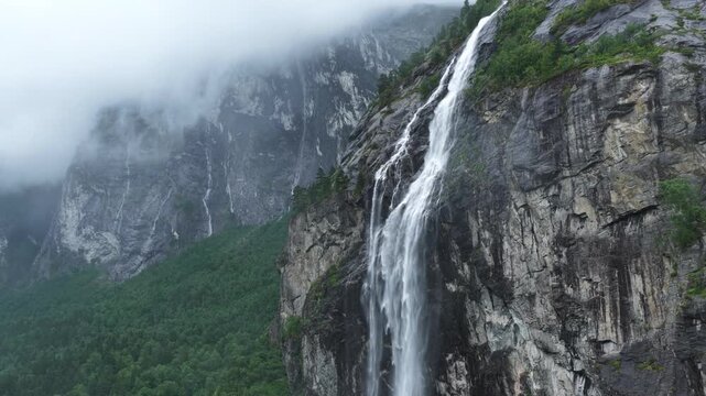 backward drone flight, View on big waterfall in Romsdalen valley, falling from a high cliff, during rainy day in summer. Steep mountain cliffs, rocks, pristine forest. Amazing wilderniss in Norway. Re