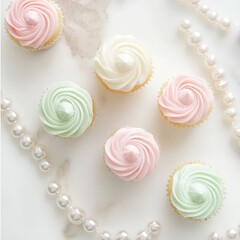 Pastel cupcakes with swirled icing and pearls arranged on a white marble surface surrounded by pearl necklaces from an overhead viewpoint
