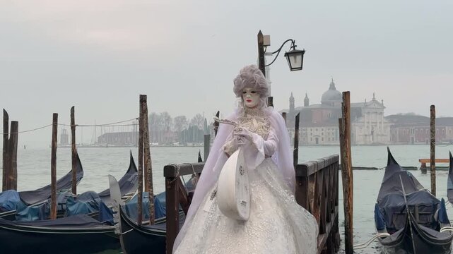 Elegant performer in white Venetian carnival costume playing a lute near gondolas on the lagoon in Venice, Italy