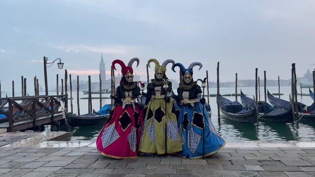 Three people dressed as Venetian carnival jesters pose near gondolas on the waterfront, wearing colorful costumes and masks against a misty lagoon backdrop