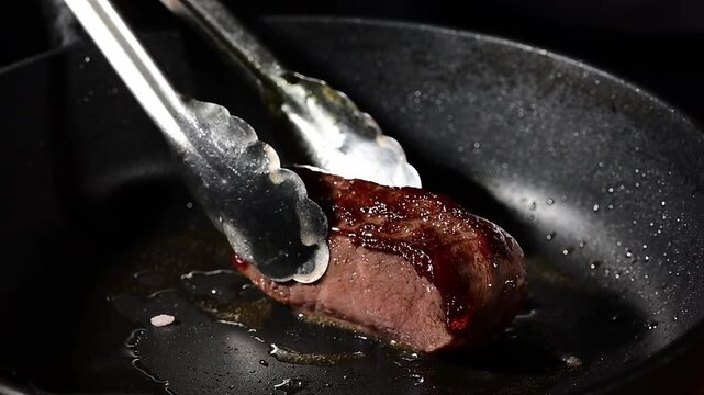 Chef flipping raw beef steak on cast iron pan with tongs, searing meat cooking process in slow motion close-up
