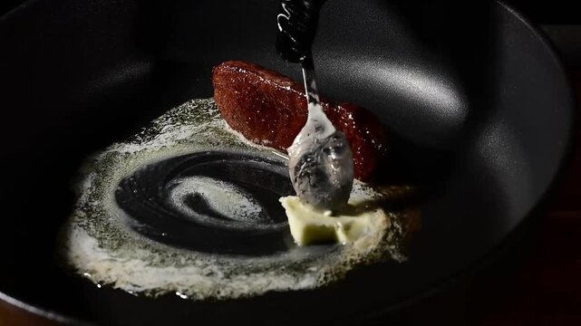 Chef basting beef steak with melted butter in cast iron skillet during searing, slow motion close-up of liquid cascading over meat surface with rising vapor