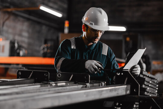 Industrial worker measuring a metal component in a factory workshop while wearing safety helmet and gloves, ensuring precision, quality control, and workplace safety in manufacturing process.