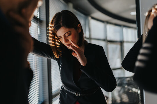 A professional woman in a black blazer leans on a window in an office. She covers part of her face with her hand, conveying worry or deep thought as light filters through blinds.