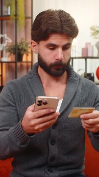 Indian man at home holds phone and bank card enters cvv completes payment gets confirmation. Arabian guy on sofa smiles satisfied looks at phone sees order confirmed relaxed mood after online purchase