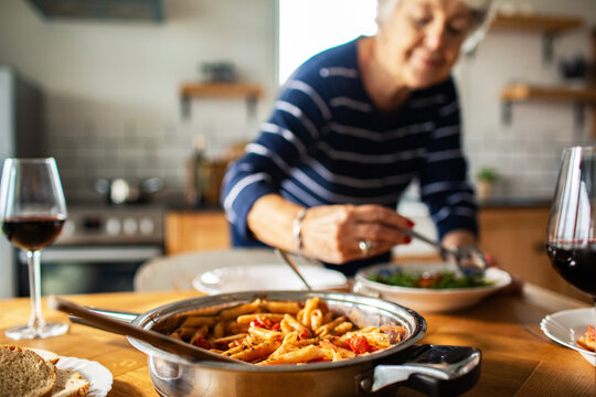 Senior woman serving pasta with wine in cozy home kitchen