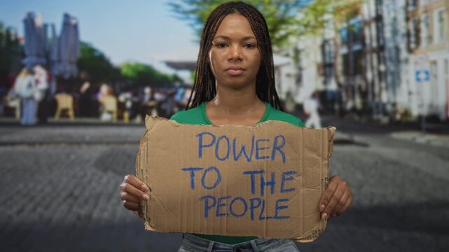 Woman holding cardboard sign on a cobblestone street in front of cafes and shops; protest empowerment determination.