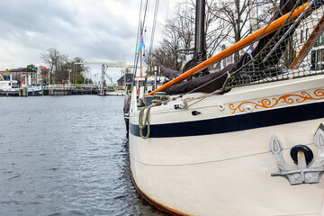 Fototapeta premium Traditional sailboat moored along canal Zwarte Water Netherlands near drawbridge village waterfront. Nautical detail rope anchor mast rigging under overcast sky creating calm maritime atmosphere