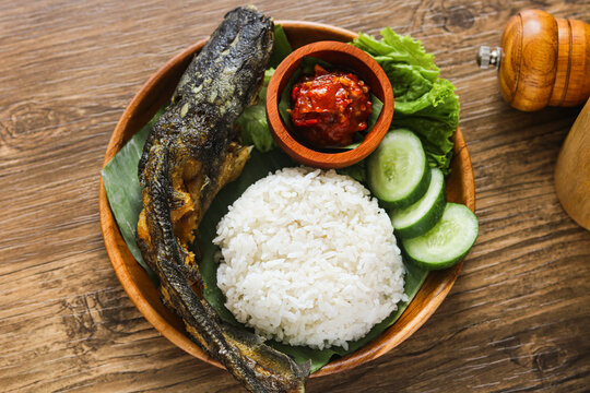 A serving of Nasi Pecel Lele, featuring deep-fried catfish paired with steamed rice, cucumber, and a signature chili sambal on a banana leaf.
