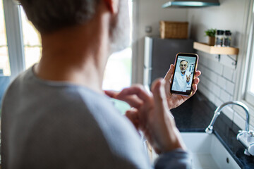 Man consulting doctor via smartphone video call at home kitchen