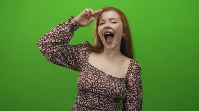 Redhead woman smiling with open mouth and raised arm gesturing at camera, bare shoulder visible in green screen studio; joyful celebration.