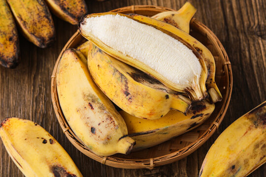 Top view of a basket filled with ripe yellow saba bananas (pisang kepok). With one fruit peeled to show its center. The wooden surface creates a warm and organic scene.