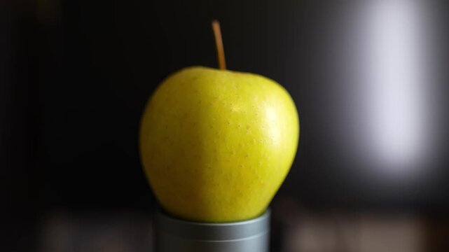 A vibrant yellow apple with a thin stem sits atop a small metallic cylindrical stand. 