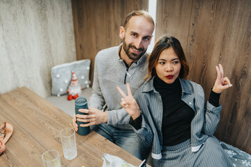 A happy couple sits at a wooden table, posing with peace signs. They enjoy a relaxed moment indoors with drinks and cozy decor.