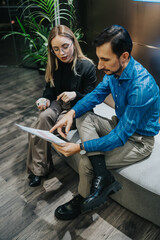 A man and a woman discuss a document in a modern office lounge. They review a report together in a professional setting.