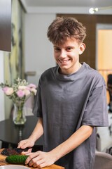 Fototapeta premium Happy teenage boy slicing cucumber in the kitchen while preparing fresh vegetable salad. Healthy lifestyle concept.