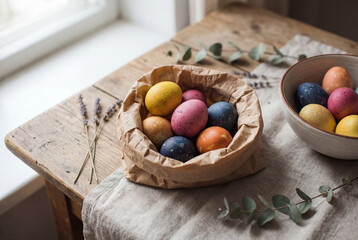 Colorful speckled Easter eggs resting in a kraft paper lined bowl on rustic wooden table with linen cloth, lavender, and greenery in soft window light