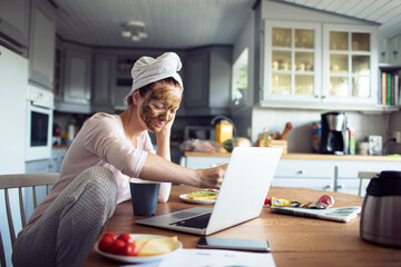 Fototapeta premium Woman with face mask working on laptop in home kitchen
