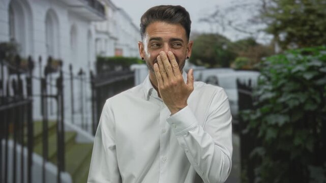 Young hispanic man covering mouth with hand on urban street by white townhouse facade; lighthearted joy.