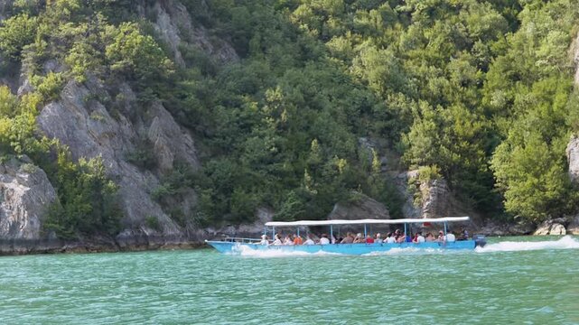 Tourist boat on turquoise Koman Lake, Albania. Lush cliffs and forest offer scenic views perfect for travel and nature documentaries.