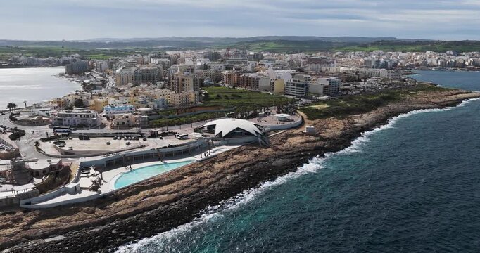Aerial view of the coast where the dark blue sea contrasts with the rocky land, and a building with a white roof stands out, St. Paul's Bay, Malta.