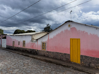 Rustic facade of a traditional pink and white colonial house with a yellow door on a cobblestone street under a melancholic sky - Lara, Venezuela