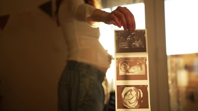 Expectant mother holding a sonogram of her unborn baby while standing at home with beautiful sunlight in the background, gently caressing her pregnant belly in a tender, loving moment