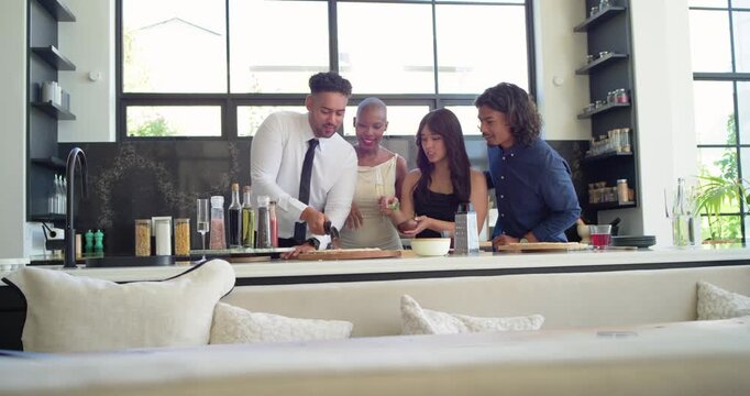 Friends preparing flatbread at kitchen island man in tie starting cut to serve, others adding herbs