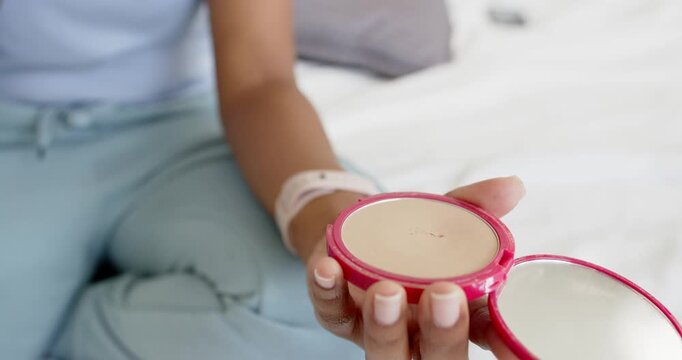 Brush is entering frame, tapping and sweeping pink compact to load bristles for makeup on bed