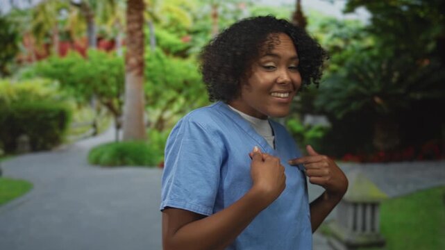 Young african american woman in blue scrubs pointing finger to chest on stone path in park; self doubt.