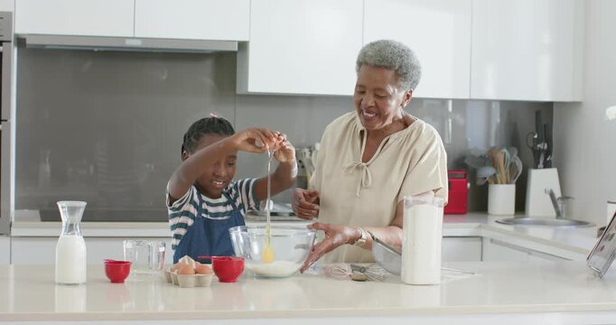 African senior woman guiding granddaughter child cracking eggs into bowl at kitchen island, baking