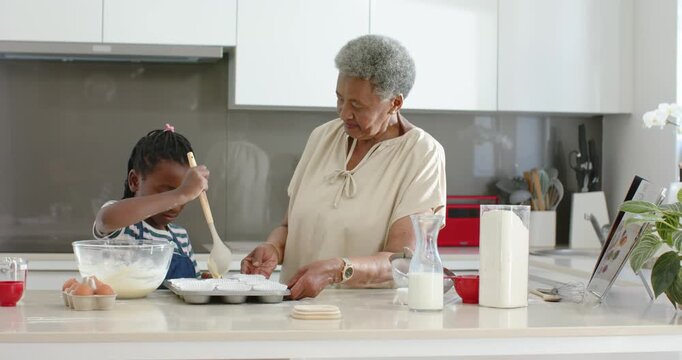 African American female teen using tablet pouring into muffin tin in kitchen while Sr holding tray