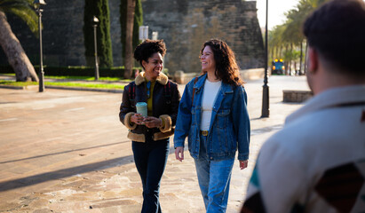 Fototapeta premium Two friends strolling a sunny city street, laughing and chatting over coffee, casually dressed and enjoying a relaxed urban weekend together in warm daylight and autumn breezes