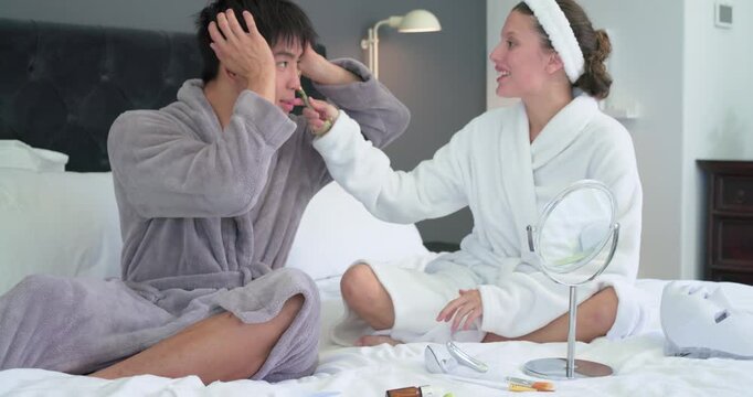 Diverse couple crosslegged on bed in robes, woman reaching and applying roller to pamper partner