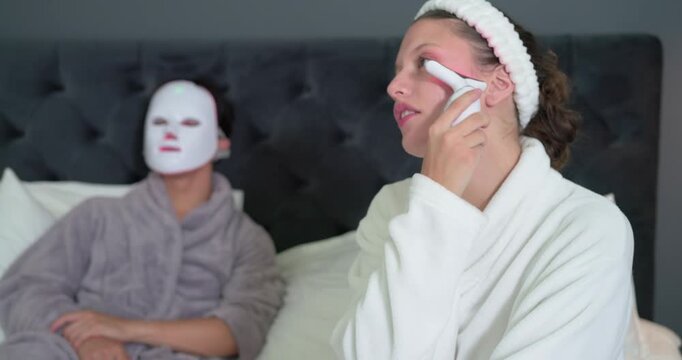 Diverse couple: woman starting skincare with device on bed while man wearing LED mask resting