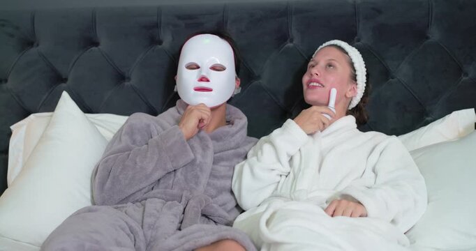 Diverse couple in robes on bed, man putting on LED mask, woman gliding device for skincare