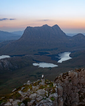 Aerial view of a solitary figure stands atop a rugged peak, overlooking a tapestry of lochs reflecting the fading light near Ullapool, Scotland, United Kingdom.