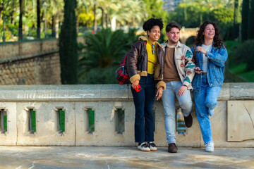 Fototapeta premium Group of young diverse friends smiling and laughing together, relaxing on a stone railing during an outdoor excursion, enjoying a sunny day in a beautiful public city park