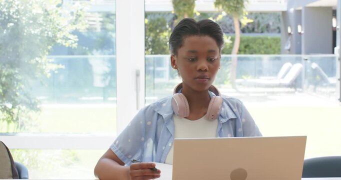 African American teen tapping pen, studying on laptop with notes, pink headset near glass door