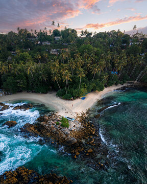 Aerial view of Secret Beach where turquoise waves crash against rocky shores, fringed by lush palm trees under a pastel sky, Mirissa, Southern Province, Sri Lanka.