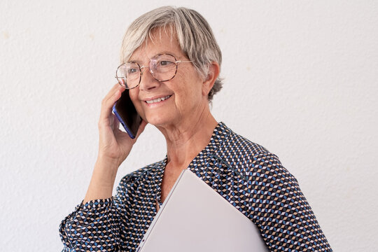 Smiling senior woman wearing round glasses standing against a neutral background using phone holding laptop under her arm. Modern elderly lady, confidence, active aging, and positive lifestyle