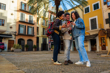 Obraz premium Three diverse friends exploring a european city square, sharing a moment and looking at their mobile devices, connecting with technology and enjoying their tourism experience together