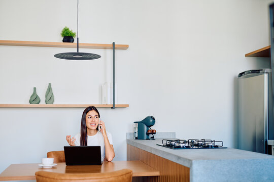 Woman talking on smartphone and working on laptop in a modern kitchen, juggling remote work with daily life