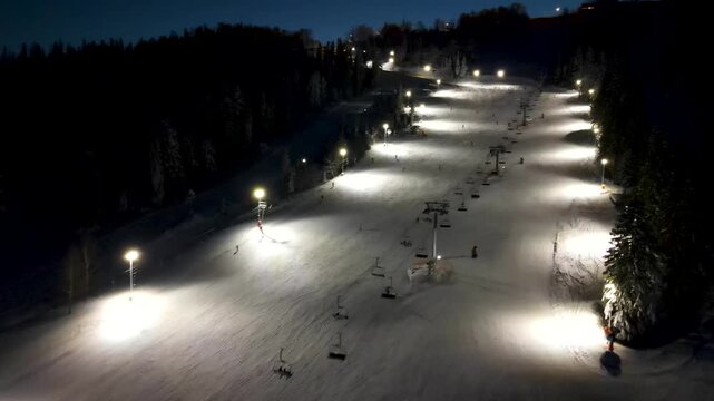 Aerial drone view of ski resort with ski slope with snow, skiers, snowboarders, pine trees and a chairlift in the mountains in winter at night. Aerial panoramic drone view from above, top view from dr
