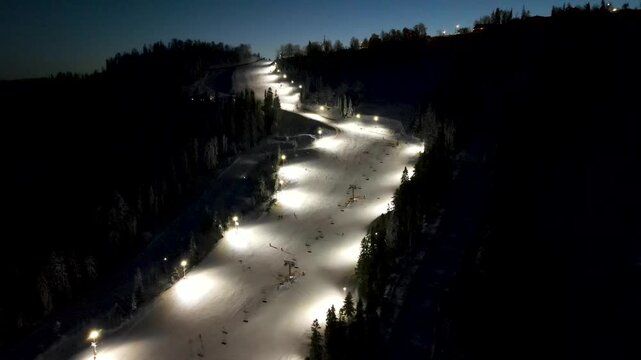 Aerial drone view of ski resort with ski slope with snow, skiers, snowboarders, pine trees and a chairlift in the mountains in winter at night. Aerial panoramic drone view from above, top view from dr