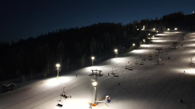 Aerial drone view of ski resort with ski slope with snow, skiers, snowboarders, pine trees and a chairlift in the mountains in winter at night. Aerial panoramic drone view from above, top view from dr