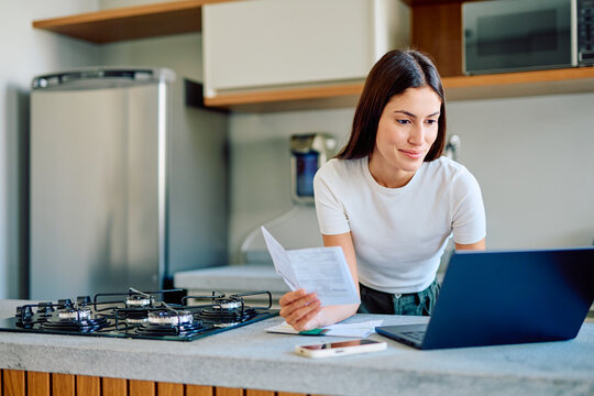 Young woman working on laptop in her kitchen, holding papers and checking bills and personal finance