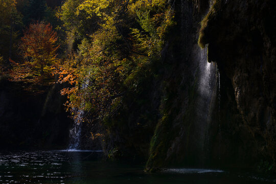 High-contrast landscape featuring waterfalls and vibrant autumn foliage against dark, moody rock faces. Professional exposure highlighting water motion and seasonal colors.